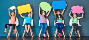 Three children sitting on chairs holding colorful speech bubbles above their heads, symbolizing communication and expression.