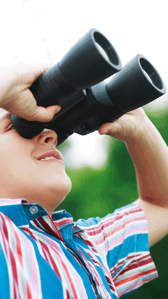 Close-up photo of a smiling child in a colorful striped shirt looking through black binoculars, with a blurred green outdoor background.