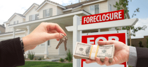 A close-up photo of a real estate transaction, showing one hand handing over house keys and another hand offering a stack of cash, with a foreclosure sign and a suburban house in the background.