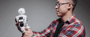 A person in a plaid shirt holding a small, white humanoid robot, both facing each other in a well-lit, neutral setting, symbolizing human and AI collaboration in digital innovation.