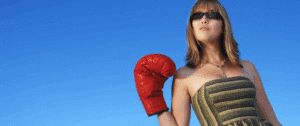 A close-up photo of a confident woman wearing sunglasses and a patterned top, holding up a bright red boxing glove against a vivid blue sky. The image conveys strength, determination, and readiness to take on challenges.