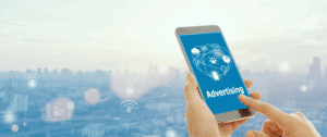 Close-up of a hand holding a smartphone with a digital advertising graphic on the screen, set against a blurred cityscape background with icons representing wireless connectivity and digital marketing.
