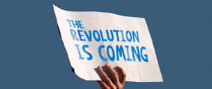 A hand holding up a white sign with blue text reading “The Revolution Is Coming” against a solid blue background, representing major changes in search behavior driven by AI, personalization, and mobile trends.