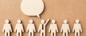 A row of wooden human figures on a brown background, with one figure raising its hand beneath a large speech bubble, symbolizing user engagement and the importance of tracking micro-conversions in digital marketing.