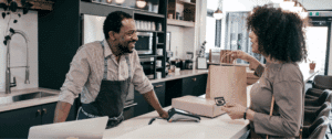 A friendly retail employee stands behind a counter, smiling and handing a shopping bag to a customer with a credit card, inside a bright, modern store. The image captures the moment of in-store purchase, representing successful conversion of website visitors into real-world shoppers.